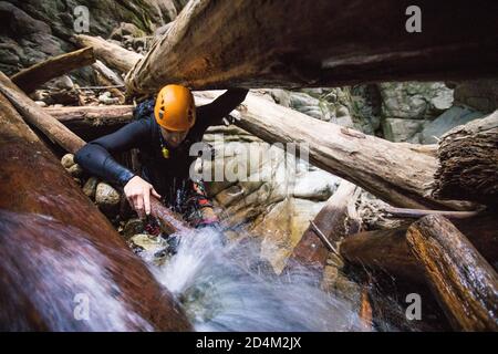 L'uomo che esplora il canyon si arrampica attraverso una confettura di tronchi, indossando casco, muta Foto Stock