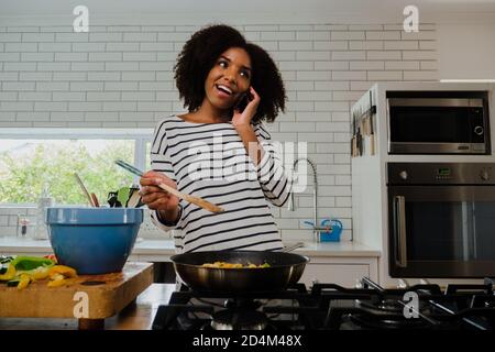 Donna che chiacchiera felicemente con il ragazzo su smartphone mentre fa una cena sana sulla stufa in cucina moderna. Foto Stock