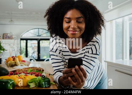 Giovane donna che naviga ricette su smartphone e sorridente mentre in cucina, ritratto Foto Stock