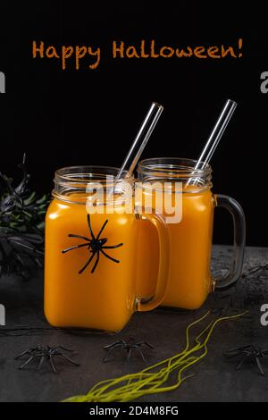 Due vasetti quadrati in vetro con coda di zucca e cannucce in vetro su un tavolo nero testurizzato decorato con ragni e piante spooky. Felice Halloween greetin Foto Stock