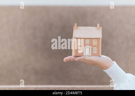 Donna mano che tiene in legno casa giocattolo in miniatura. Costruzione casa, bene immobile e concetto di proprietà. Foto Stock
