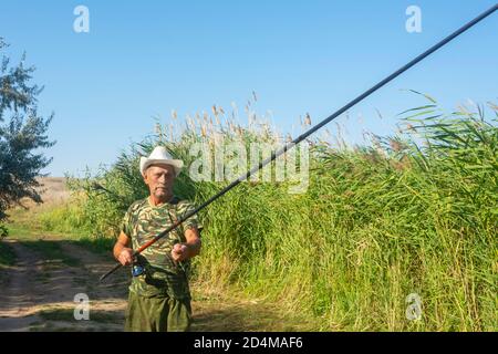 Un vecchio pescatore prepara canna da pesca in estate sulla riva del fiume in canne. Un uomo anziano in un cappello bianco ama la pesca sportiva. Sole luminoso, messa a fuoco morbida Foto Stock