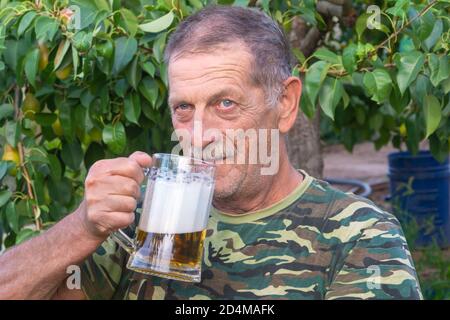 L'uomo anziano con baffi trattiene la tazza di birra leggera con la schiuma. Il pensionato riposa all'aperto. Concetto di relax e alcol, concentrazione selettiva Foto Stock