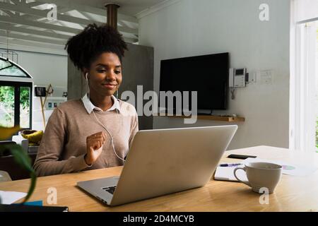 Sorridente donna africana con auricolari che indossano afro mentre si sta facendo una videochiamata con colleghi di lavoro. Seduto al tavolo da cucina con caffè caldo Foto Stock