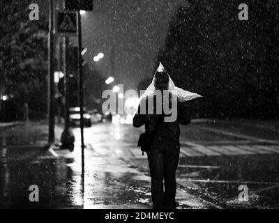 Attraversamento pedonale in città di notte durante la pioggia intensa. Silhouette di un uomo con una borsa di plastica sulla testa invece di un ombrello. Fari per auto Foto Stock