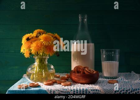Dandelioni e latte. Vita still rustica. Bouquet di dandelioni, latte in bottiglia e bagel. Sfondo di legno verde. Foto Stock