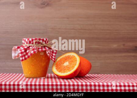 Marmellata d'arancia in vaso di vetro e metà arancione su una tovaglia a scacchi rossi. Sfondo di legno Foto Stock