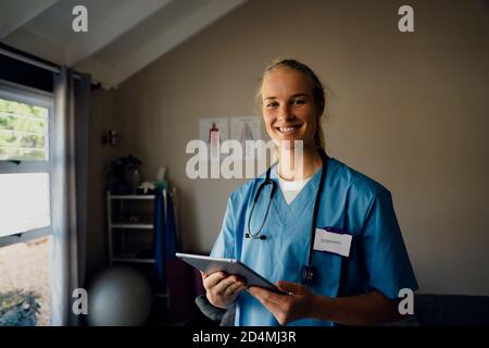 Ritratto di giovane bella dottoressa caucasica in scrub con tavoletta digitale, sorridente Foto Stock