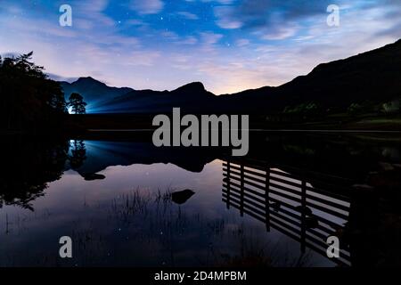 Un colpo di notte nuvoloso di Blea Tarn in inglese Lake District con auto fari che riflettono sul Tarn Foto Stock