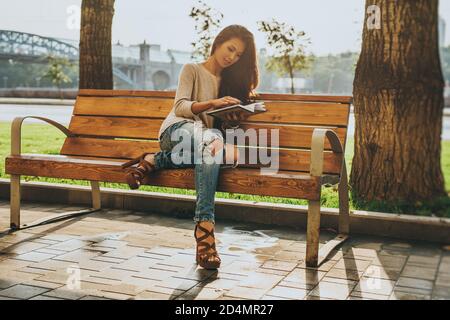 Giovane donna asiatica di moda e bellezza seduta su panca dentro città e libro di lettura Foto Stock