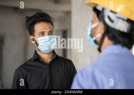 Spalla colpo, lavoratore di costruzione e ingegnere in loco parlando indossando maschera medica a distanza sociale - concetto di business, settore riaprire Foto Stock