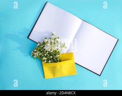 Busta di carta gialla con fiori di camomilla bianchi da giardino e blocco note vuoto aperto su sfondo azzurro. Modello floreale festivo. Saluto c Foto Stock