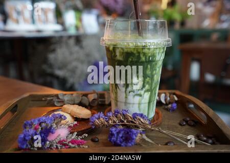 Primo piano take away in plastica di Matcha verde ghiacciato tè al latte servito con biscotti al burro su tavola di legno decorata con fiori secchi e chicchi di caffè Foto Stock