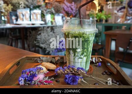 Primo piano take away in plastica di Matcha verde ghiacciato tè al latte servito con biscotti al burro su tavola di legno decorata con fiori secchi e chicchi di caffè Foto Stock