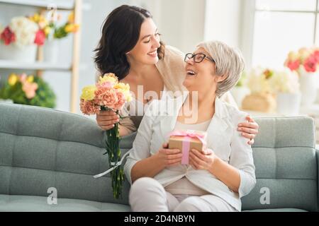 Buon giorno della madre! Bella giovane donna e sua madre con fiori e confezione regalo a casa. Foto Stock