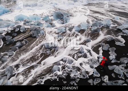 Uomo vestito rosso che fotografa le onde tra le rocce di ghiaccio nella famosa spiaggia nera islandese. Spiaggia nera, ghiaccio strega diamanti paesaggio al vostro calendario Foto Stock