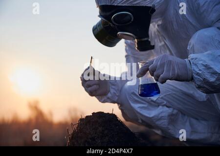 Vista laterale, immagine ravvicinata di un ecologo accoccolato che indossa una tuta bianca, una maschera a gas e guanti che raccolgono campioni di antrillo bruciato, tenendo un pallone di vetro con liquido blu e pinzette al tramonto Foto Stock