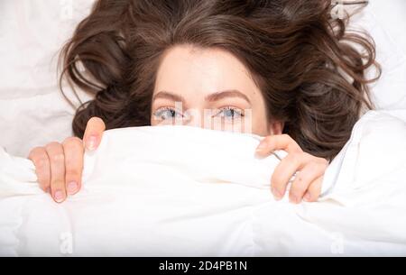 Closeup di bella giovane donna con i capelli lunghi che si stende e si nasconde sotto coperta bianca Foto Stock