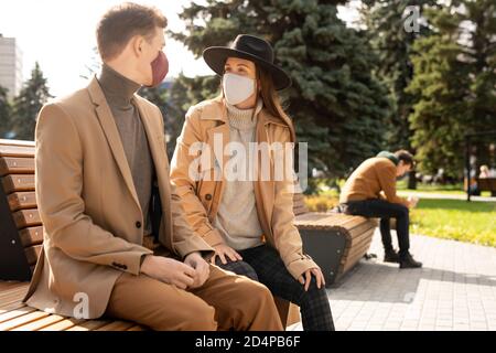 Felice giovane coppia elegante in maschere protettive rilassandosi sulla panca nel parco Foto Stock