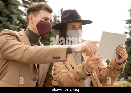 Giovane coppia elegante in maschere protettive e casualwear elegante riposo nel parco Foto Stock