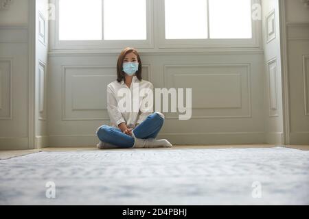 giovane donna asiatica in quarantena a casa con maschera facciale seduta sulle gambe del pavimento incrociate Foto Stock