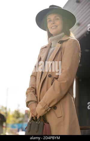 Bella donna con cappello nero, trench beige e pullover grigio che ti guarda Foto Stock