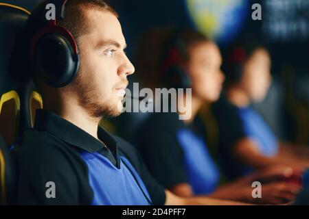 Vista laterale di un ragazzo caucasico focalizzato, giocatore di cybersport maschile indossando cuffie che gioca online video giochi in team professionale, partecipando al torneo eSport mentre si siede nel club di gioco Foto Stock