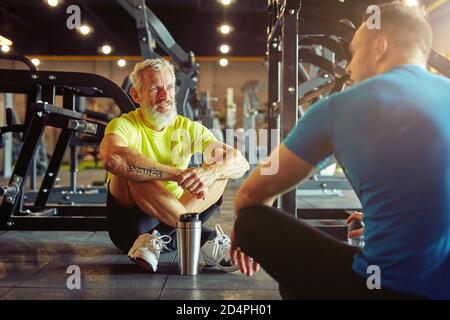 Riposo dopo l'allenamento. Atletico uomo di mezza età discutere qualcosa con istruttore di fitness o personal trainer mentre si siede insieme sul pavimento Foto Stock
