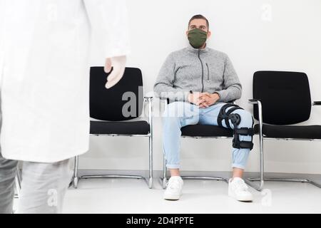 Paziente giovane con maschera facciale in una sala d'attesa bianca di un ospedale che guarda un medico Foto Stock