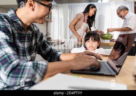 Ragazza asiatica sconvolto che guarda e aspetta suo padre che lavora da casa, mentre la città si trova nella sala da pranzo, mentre mamma e nonno cucina. Dom Foto Stock