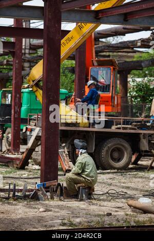 Saldatore in tuta protettiva e maschera in primo piano. Gru e operatore gru sullo sfondo sfocati. Costruzione di edifici industriali. Foto Stock