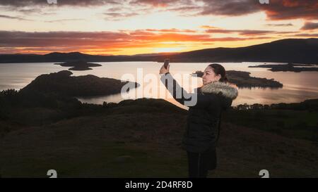 Una donna che fa un selfie da uno smartphone si trova su una montagna vicino al lago sullo sfondo di un cielo di tramonto ardente. Conic Hill, Scozia, United Foto Stock
