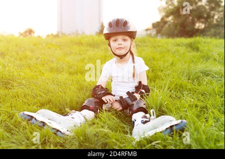 Bambina carina su pattini a rotelle in casco seduto l'erba al parco Foto Stock