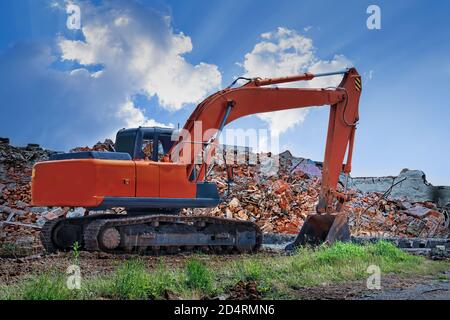 Un escavatore arancione si trova sullo sfondo di un edificio distrutto e di un cielo blu. Foto Stock