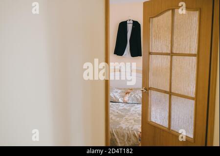 Vista attraverso la porta aperta bella tuta nera camicia dello sposo bianco appeso su un appendiabiti. Foto Stock
