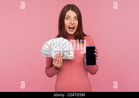 Donna bruna eccitata estremamente sorpresa in felpa rosa che tiene smartphone con schermo vuoto e bollette del dollaro guardando la fotocamera con shock. Interno Foto Stock