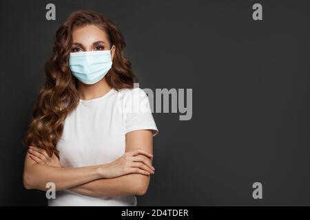 Donna sicura che indossa una maschera medica protettiva e una t-shirt bianca in piedi con braccia incrociate su sfondo nero Foto Stock