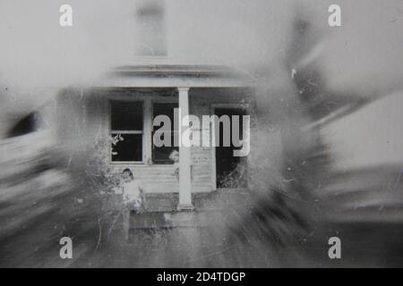 Bella fotografia in bianco e nero d'epoca degli anni '70 di una vecchia casa di famiglia in un sogno con una bambina in piedi davanti ad essa. Foto Stock