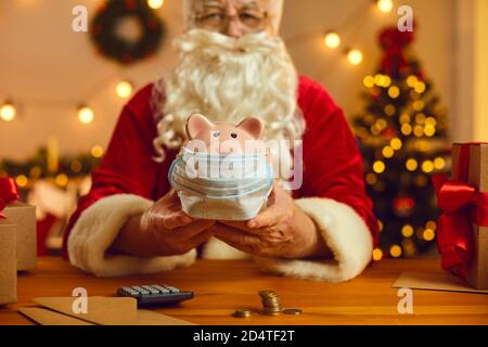 Babbo Natale alla scrivania con soldi e calcolatrice che tiene il piggy banca che indossa una maschera medica Foto Stock