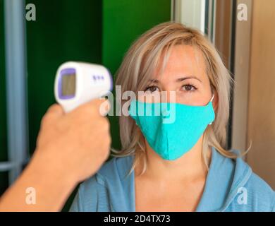 Una donna in una maschera medica viene misurata la temperatura mediante un termometro elettronico a infrarossi all'ingresso dell'ufficio. Sembra stanca e malata. Mettere a fuoco il viso. Foto Stock