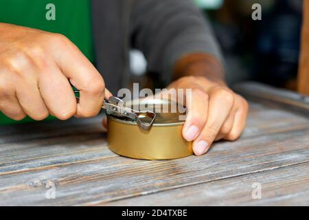 Le mani dell'uomo che aprono una lattina su un vecchio tavolo di legno con un vecchio apriballe con un manico di metallo. Cibo in scatola e un coltello speciale. Profondità di campo poco profonda. Foto Stock