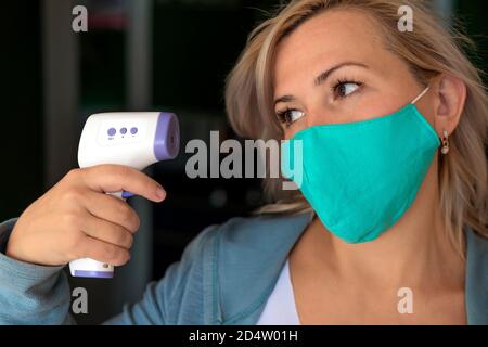 La donna bionda punta un termometro a infrarossi al suo viso come se fosse una pistola e lo guarda. Indossa una maschera medica. Self care, concetto medico e divertente. Foto Stock
