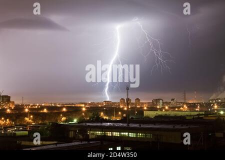 Un potente fulmine colpisce la città di notte. Un forte colpo di fulmine su un cielo grigio scuro colpisce il terreno, illuminando l'area industriale A. Foto Stock