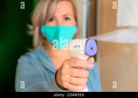 Misurazione della temperatura mediante termometro elettronico a infrarossi. Concetto di prevenire l'infezione, focolaio di malattia di Coronavirus COVID-19. Donna caucasica in maschera medica con termometro. Concentrarsi sui dispositivi elettronici. Profondità di campo poco profonda. Foto Stock
