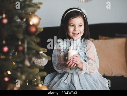 Sorridente bambina di 4-5 anni seduta sotto l'albero di Natale con decorazioni in camera primo piano. Anno nuovo. Stagione invernale. Foto Stock