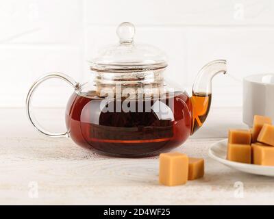 Teiera in vetro trasparente piena di tè nero caldo appena preparato vicino alla tazza e caramello alla vaniglia su un tavolo di legno bianco. Tè bevendo con dolci. Vista frontale Foto Stock