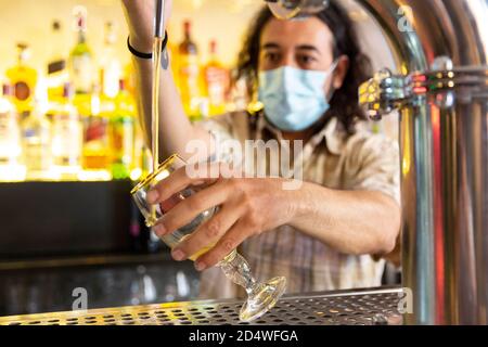 Primo piano di un barman che indossa una maschera medica che riempie un bicchiere di birra in un bar moderno. Concetto di nuove restrizioni nella vita notturna. Messa a fuoco selettiva. Foto Stock