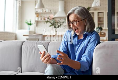 Stupita donna anziana matura che legge il messaggio sullo smartphone a casa. Foto Stock