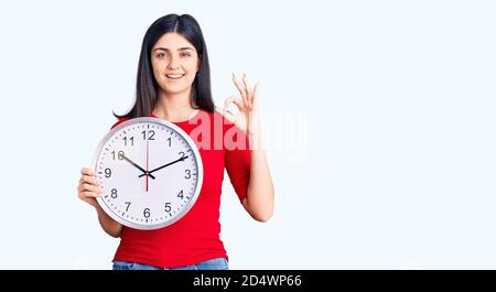 Giovane bella ragazza tenendo orologio fare ok segno con le dita, sorridente amichevole gesturing eccellente simbolo Foto Stock