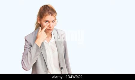 Giovane bella donna bionda che indossa una giacca elegante che punta all'occhio che ti guarda gesto, espressione sospetta Foto Stock